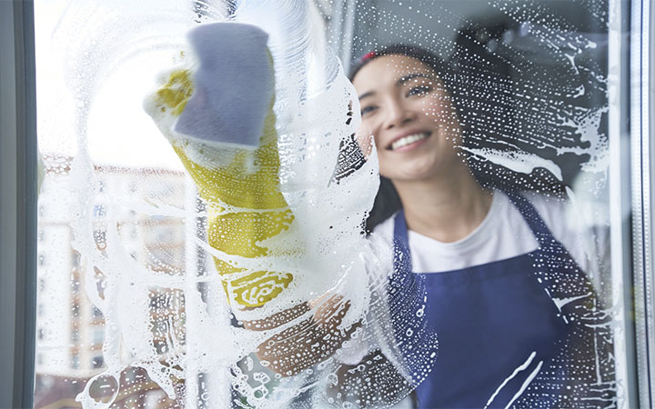 limpeza_0005_mulher-jovem-alegre-sorrindo-enquanto-limpa-a-janela-a-superficie-de-vidro-usando-uma-esponja-trabalho-dome